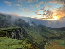 Weitläufige, grün bewachsene bergige Landschaft, über der Sonnenstrahlen aus einer Wolkendecke hervorkommen